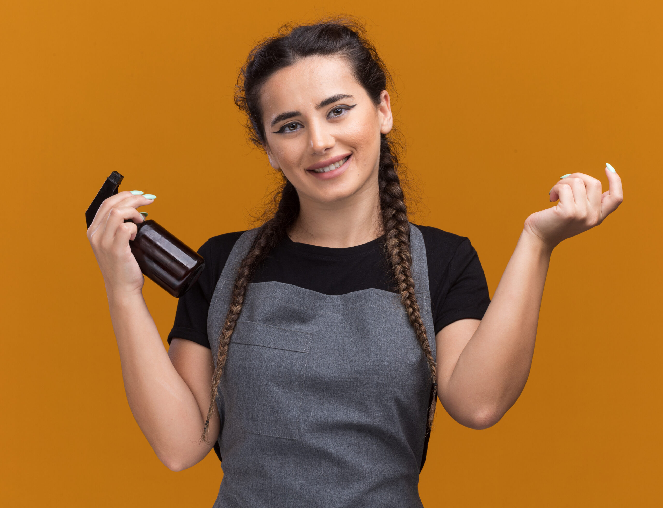 smiling young female barber uniform holding spray bottle spreading hand isolated orange wall scaled